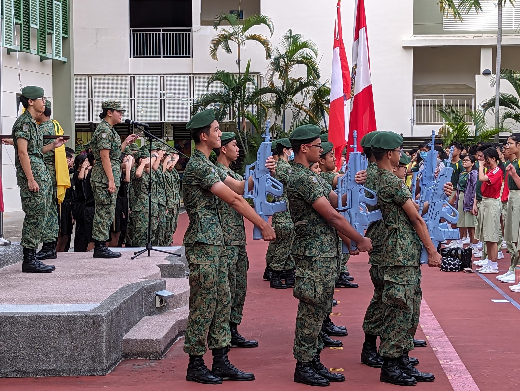 Young cadets in green camouflage uniforms holding blue rifles stand in formation during a school parade ceremony.